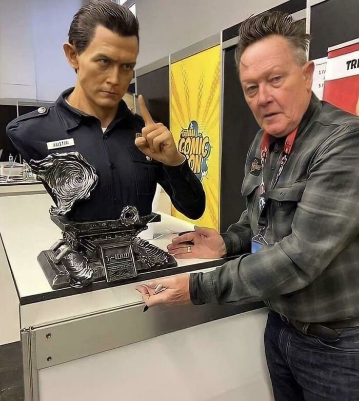 "Man posing with a futuristic police officer statue at a comic convention, highlighting funny aspects of getting old."