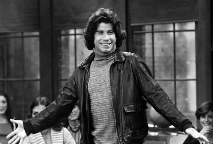 A man with curly hair in a leather jacket smiling, embodying humor about getting old faster than expected.