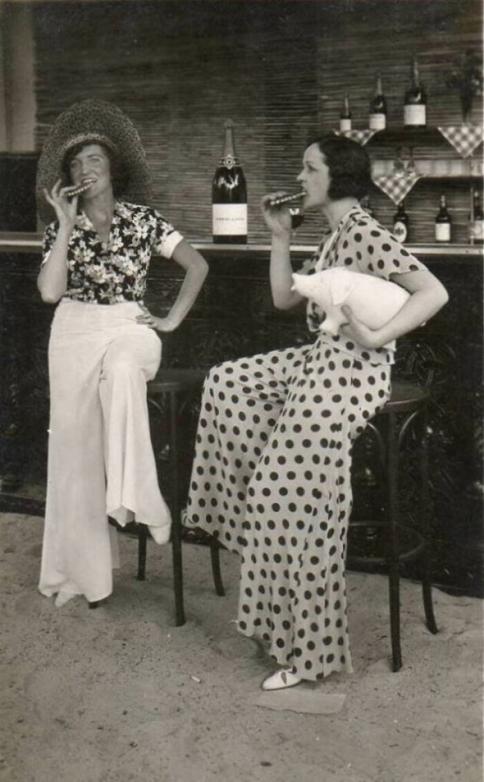 Two women in vintage outfits sitting on stools at a bar, one holding a pig figurine, in a historical photo.