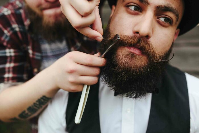Barber shaving a bearded man with a straight razor in a vintage setting, representing useless facts to impress friends