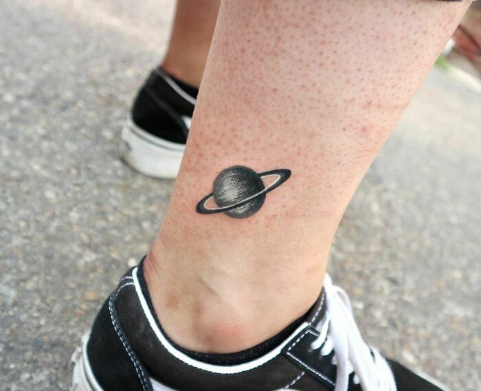 Ankle tattoo of a planet design with black sneakers in the background on a paved surface.