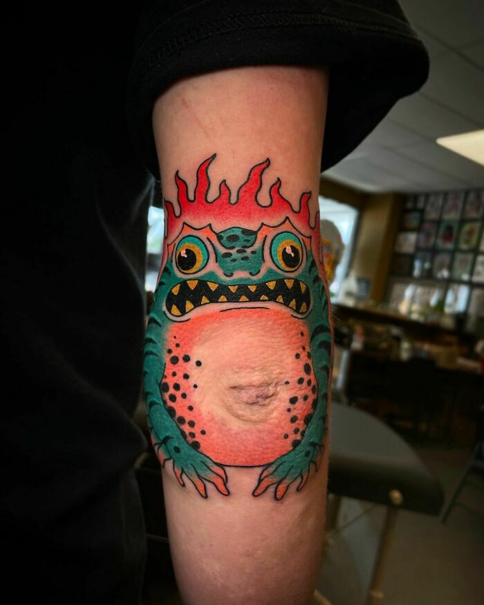 Colorful frog-themed elbow tattoo with flames in a tattoo studio setting.