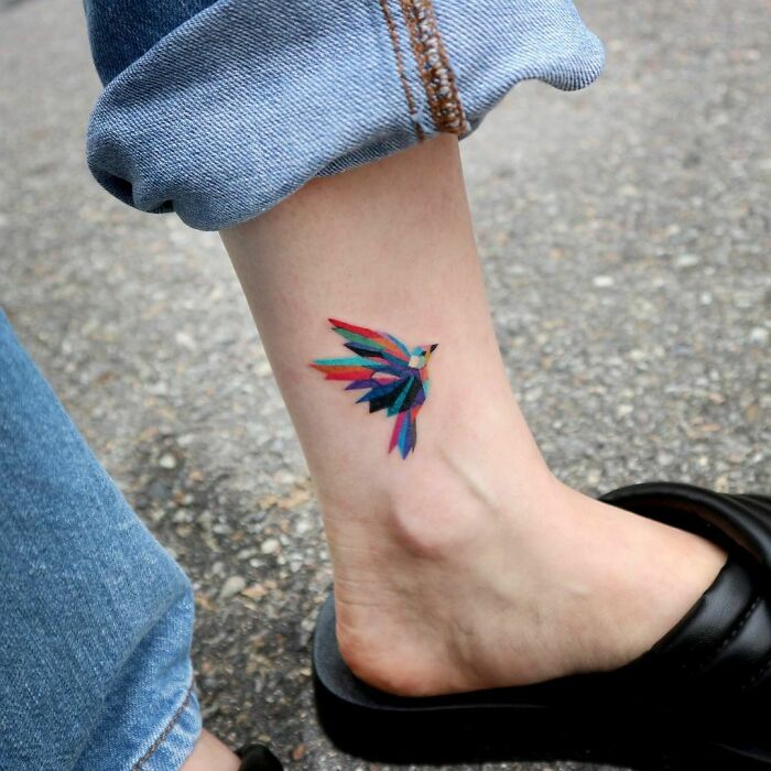 Colorful bird ankle tattoo on woman wearing jeans and black sandal.