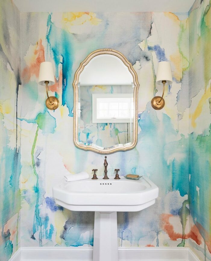 Bathroom with trendy watercolor wallpaper, white pedestal sink, ornate mirror, and gold wall sconces in a stylish space.