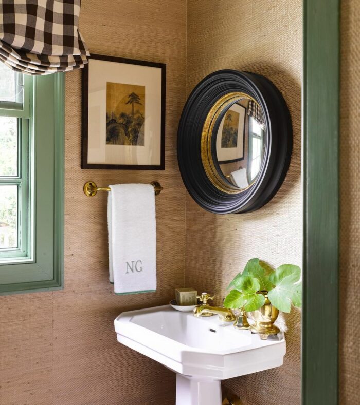 Bathroom with brown grasscloth wallpaper, black round mirror, white pedestal sink, and gold fixtures in a stylish space.
