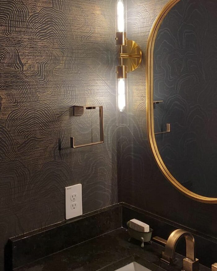 Bathroom interior featuring black striped metallic wallpaper, gold fixtures, and a round mirror above the sink.