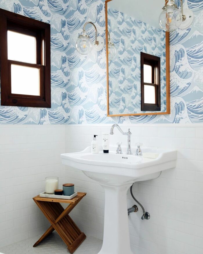 Bathroom with blue ocean wallpaper, white pedestal sink, wooden window frame, and small wooden side table with candles.