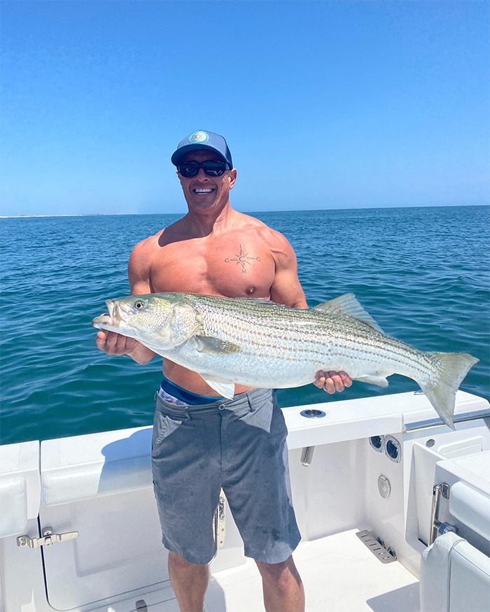 Shirtless buff celebrity holding a large fish on a boat with sunglasses and a cap against a clear blue ocean background.