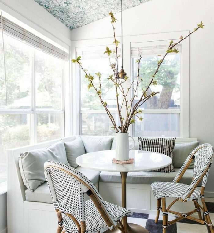 White banquette seating with gray pillows and a round table by windows, featuring cozy banquette seating ideas.
