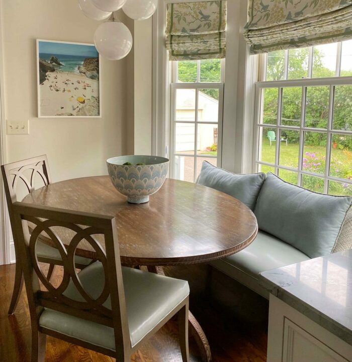 Light green banquette seating with wooden round table and chairs by a window overlooking a garden.
