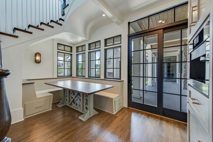 White banquette seating under stairs with wooden table in bright dining nook featuring large windows and glass doors.