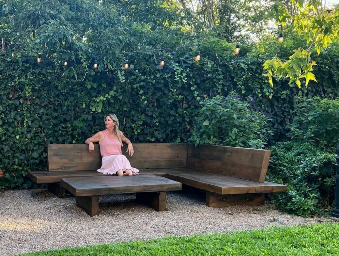 Woman sitting on a wooden banquette in a lush garden, showcasing cozy banquette seating ideas outdoors.