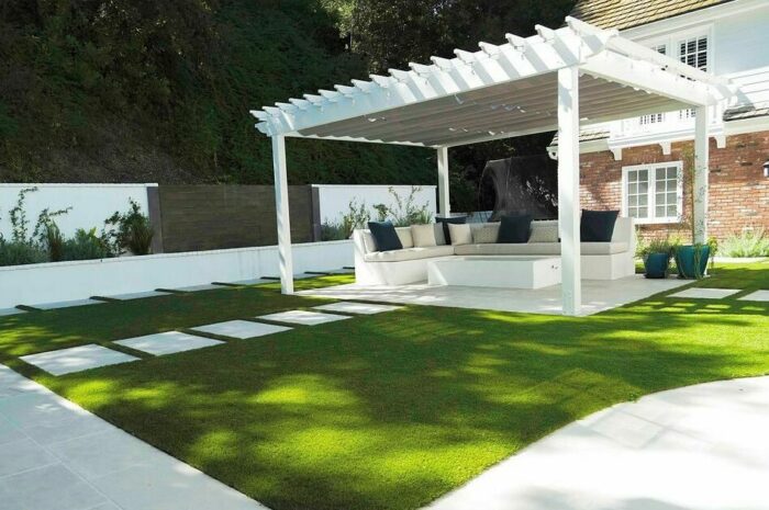 White backyard banquette seating under a white pergola surrounded by green grass and modern landscaping.