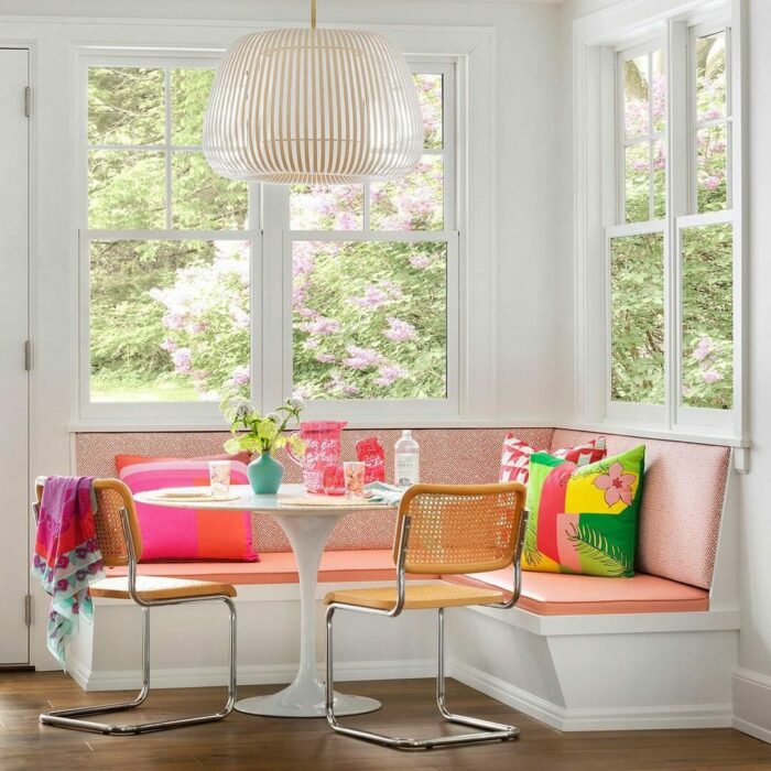 White pink banquette seating with colorful pillows around a round table and modern chairs by bright windows.