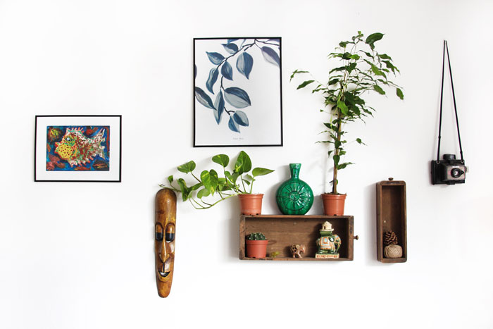 White wall decorated with boho decor featuring paintings, a wooden mask, plants, vase, and hanging vintage camera.