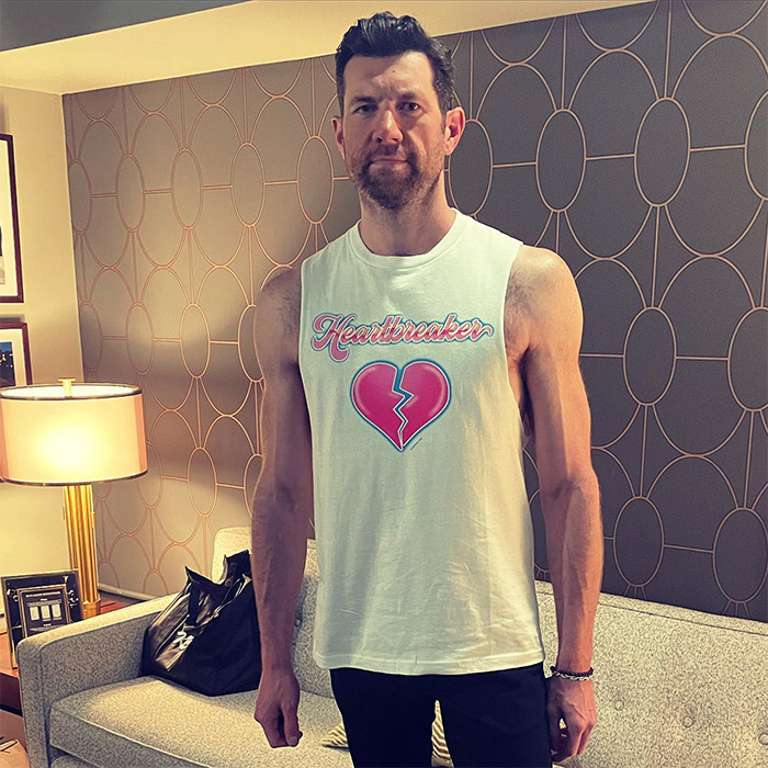 Man showing muscular arms wearing a sleeveless shirt that says Heartbreaker in a stylish living room setting.