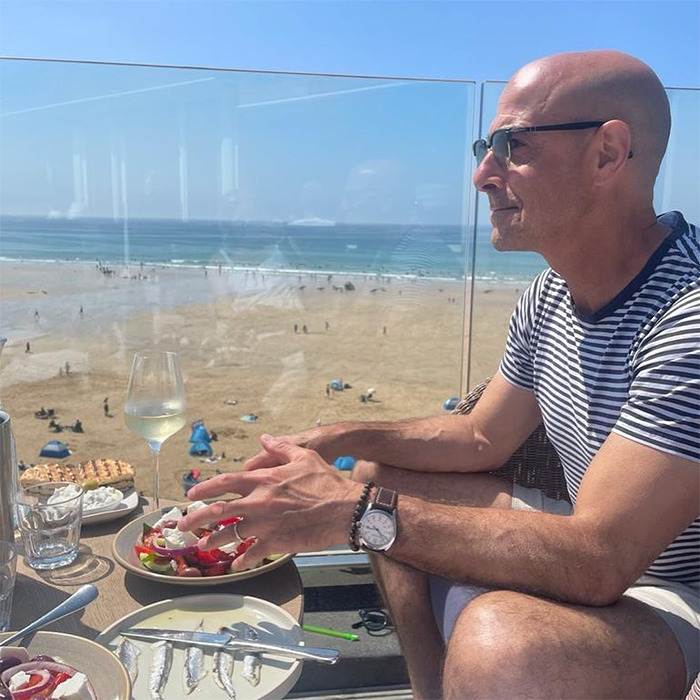 Man with muscular arms wearing striped shirt, enjoying meal and drink at seaside restaurant with ocean view.