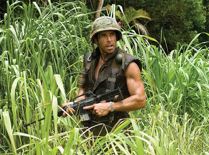 Actor in military gear showing off buff arm muscles while walking through tall grass in a jungle setting.