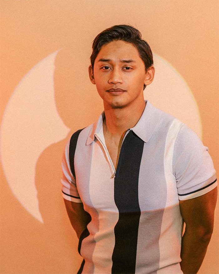 Young man in a striped polo shirt posing against an orange backdrop with a circular light, showcasing a buff appearance
