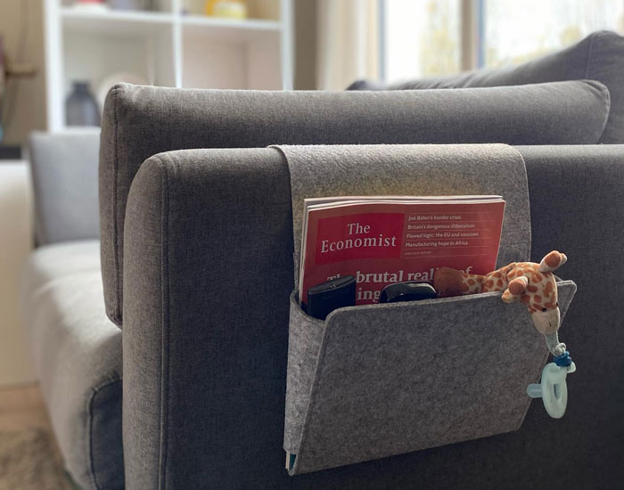 Felt side pocket attached to a grey cheap couch holding magazines, remotes, and a toy in a bright living room setting. Felt side pocket attached to a grey cheap couch holding magazines, remotes, and a toy in a bright living room setting.