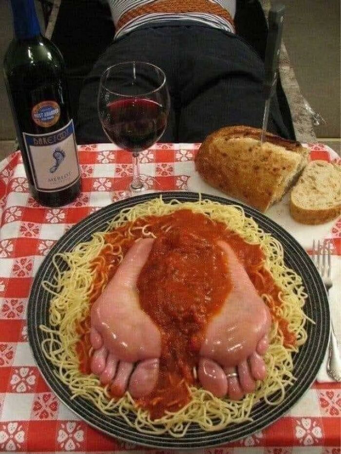 Cursed food featuring feet-shaped meat atop pasta with tomato sauce, alongside bread and red wine on a checkered tablecloth.