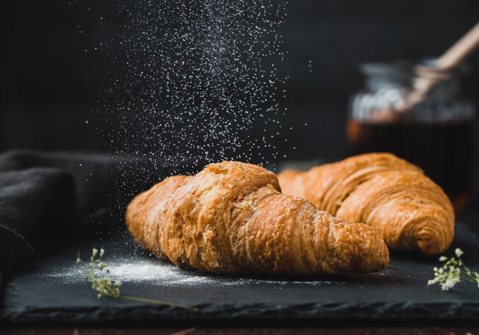 Two brown croissants on a dark surface with powdered sugar falling, showcasing culture facts reflecting diversity and beauty.