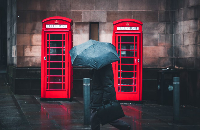 Person walking with umbrella beside iconic red telephone booths in Manchester, showcasing culture diversity and beauty of the world.