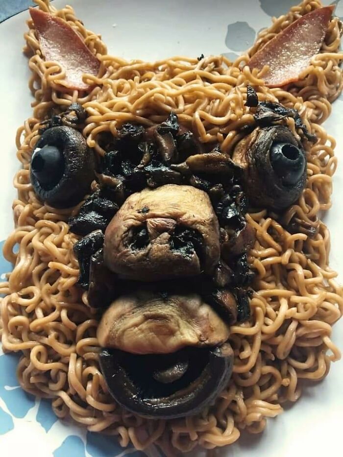 Cursed food art made with ramen, mushrooms, and bacon forming an animal-like face on a plate.