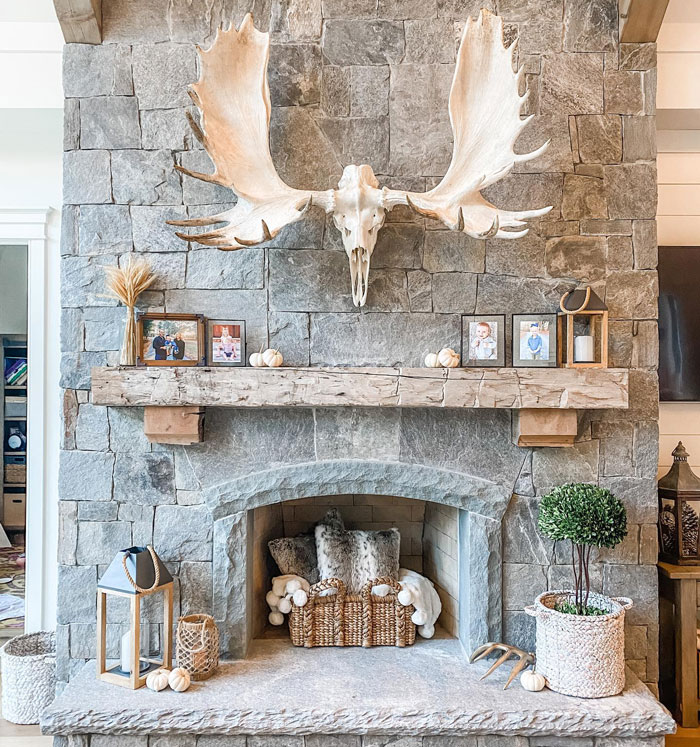 Stone living room fireplace mantel with rustic wood beam, moose antlers, and cozy decorative accents.