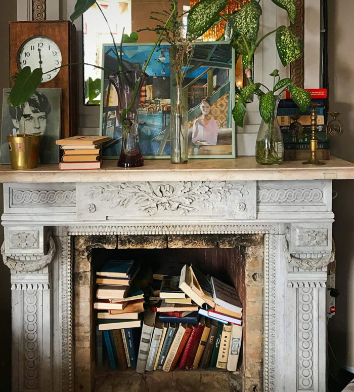 Antique fireplace mantel filled with books inside, topped with plants, artwork, and decorative items for a cozy hearth.