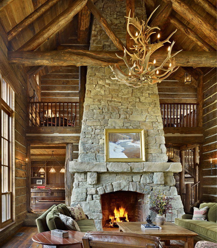 Large stone fireplace with big stone mantel in rustic living room featuring antler chandelier and cozy seating area.