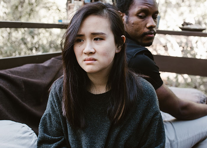 Young Asian woman and Black man sitting back to back outdoors, showing signs that inevitably make people look older.
