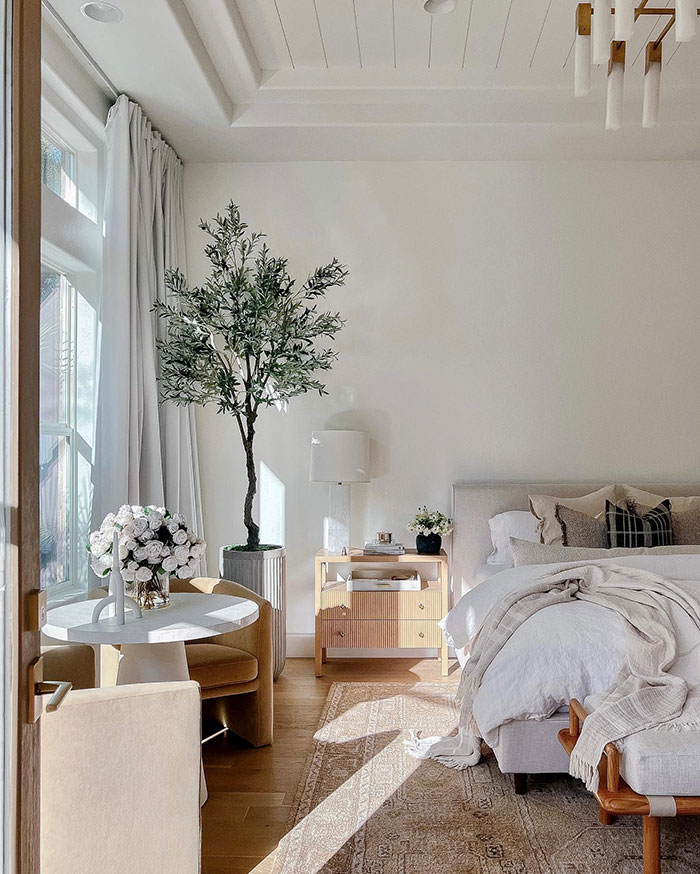Spacious bright master bedroom with big windows, neutral decor, indoor plants, cozy seating, and soft natural lighting.