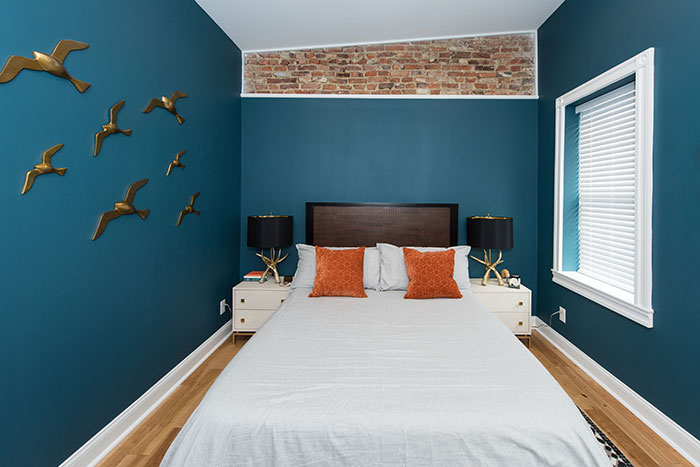 Small blue master bedroom with brick accent wall, white bedding, orange pillows, and gold bird wall decor in a cozy design.