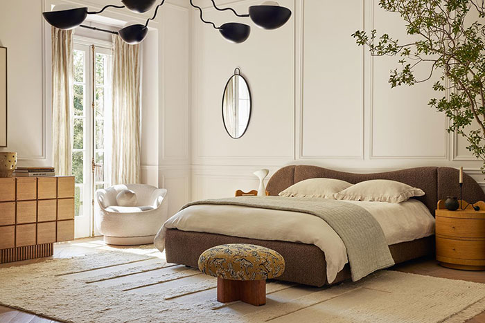 Spacious master bedroom featuring a brown upholstered bed, soft neutral tones, modern lighting, and natural light from large windows.