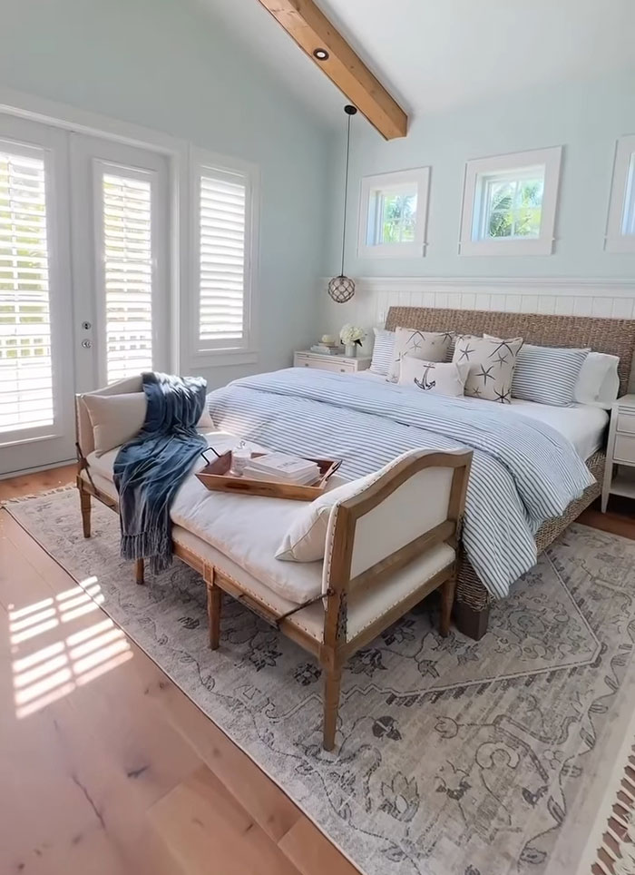 Spacious bright master bedroom featuring a striped bed, cushioned bench, and natural light through white shutters.