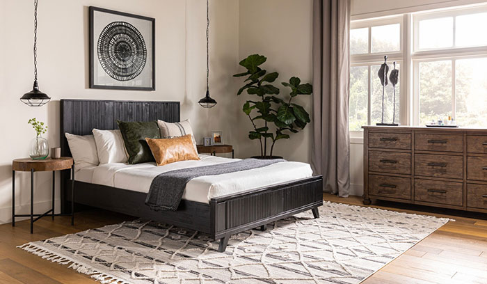 Spacious cozy rustic master bedroom with wooden bed frame, patterned rug, large dresser, and natural light from tall windows.
