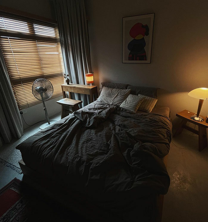Cozy master bedroom design with warm lighting, wooden furniture, a standing fan, and neutral-toned bedding and curtains.