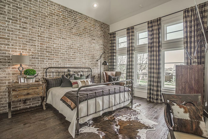 Cozy master bedroom design featuring an exposed brick wall, rustic metal bed frame, and natural light from large windows.