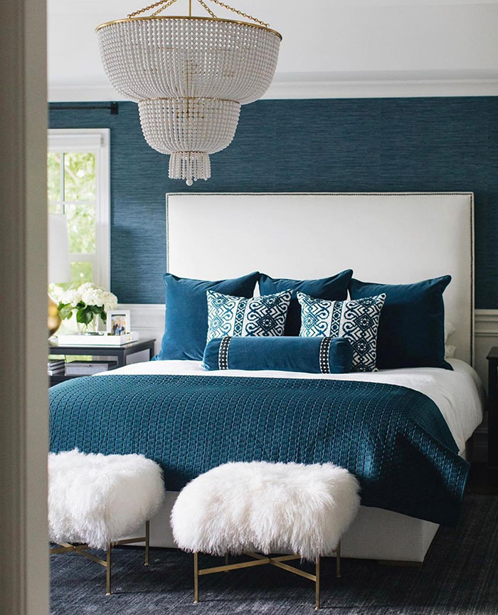Cozy blue and white master bedroom with patterned pillows, textured blanket, fluffy stools, and elegant chandelier design ideas.