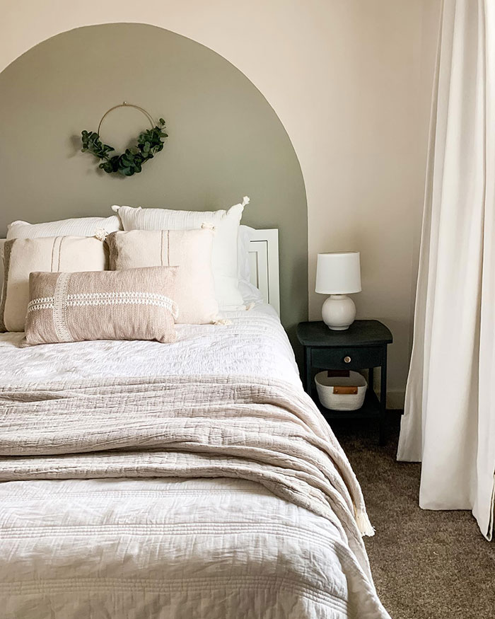 Cozy master bedroom design with neutral bedding, a white painted headboard, and a leafy wreath on soft green wall.
