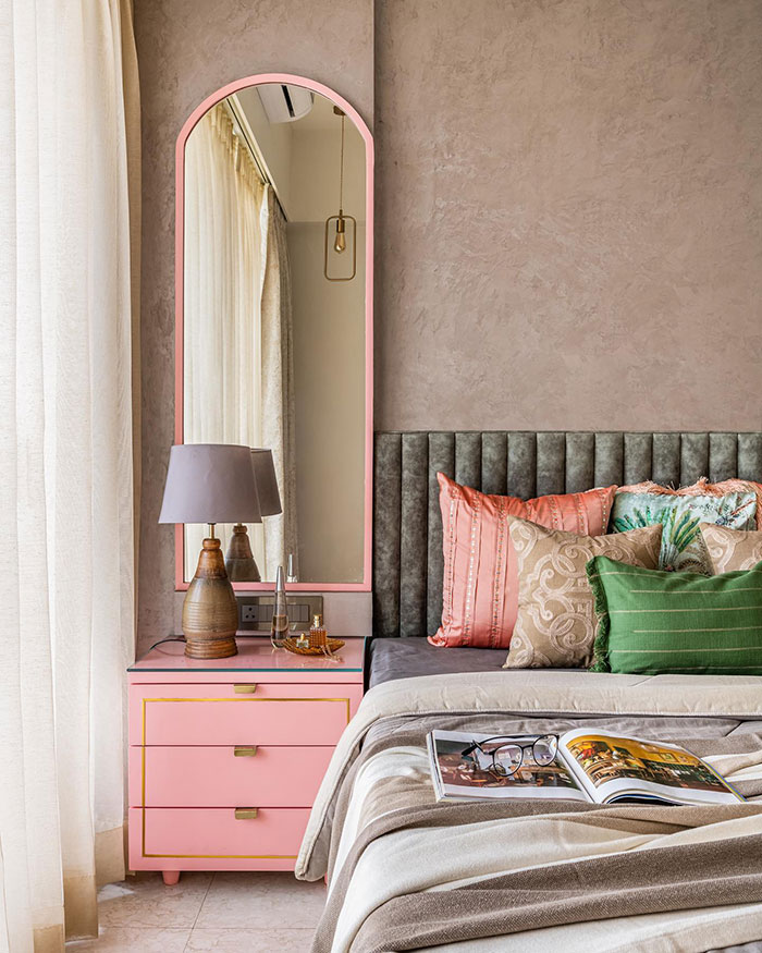Vibrant colored master bedroom with pink nightstand, green and peach pillows, gray headboard, and a tall mirror by the window.