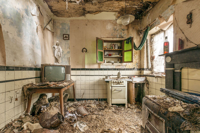 Abandoned post-apocalyptic kitchen with decayed furniture, broken appliances, and debris covering the floor and surfaces.