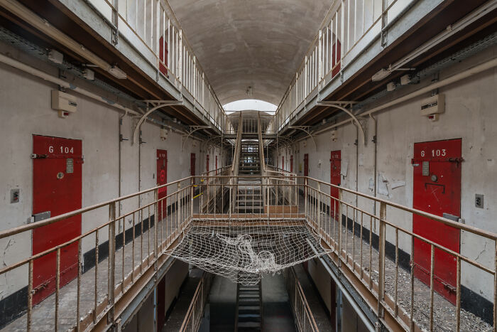 Interior of an abandoned post-apocalyptic prison corridor with rusted railings and red cell doors on both sides.