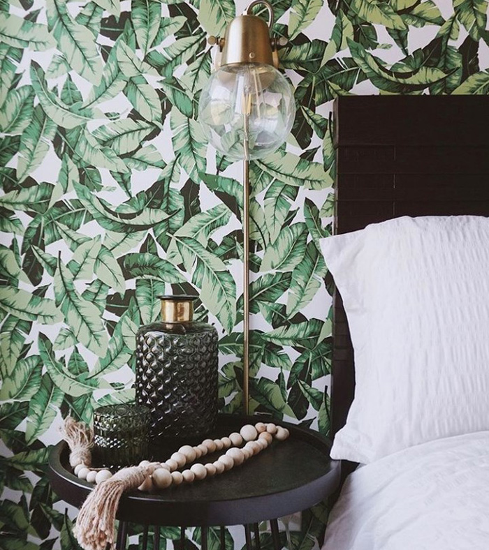 Bedroom with green leaves removable wallpaper, a dark side table, decorative bottles, and a white pillow by the headboard.