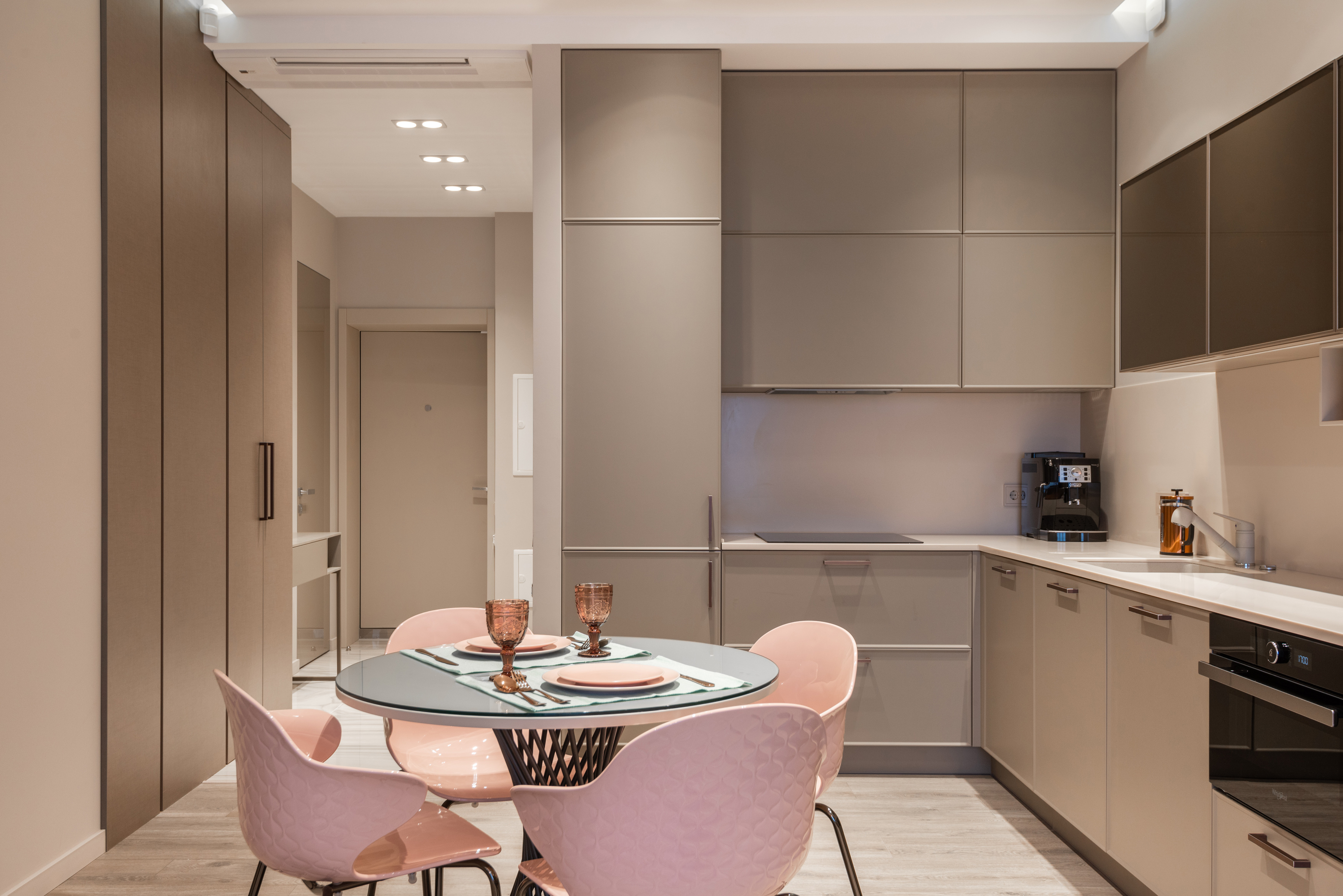 Modern kitchen with taupe cabinets, pink chairs, and a round glass table set with plates and amber glasses.