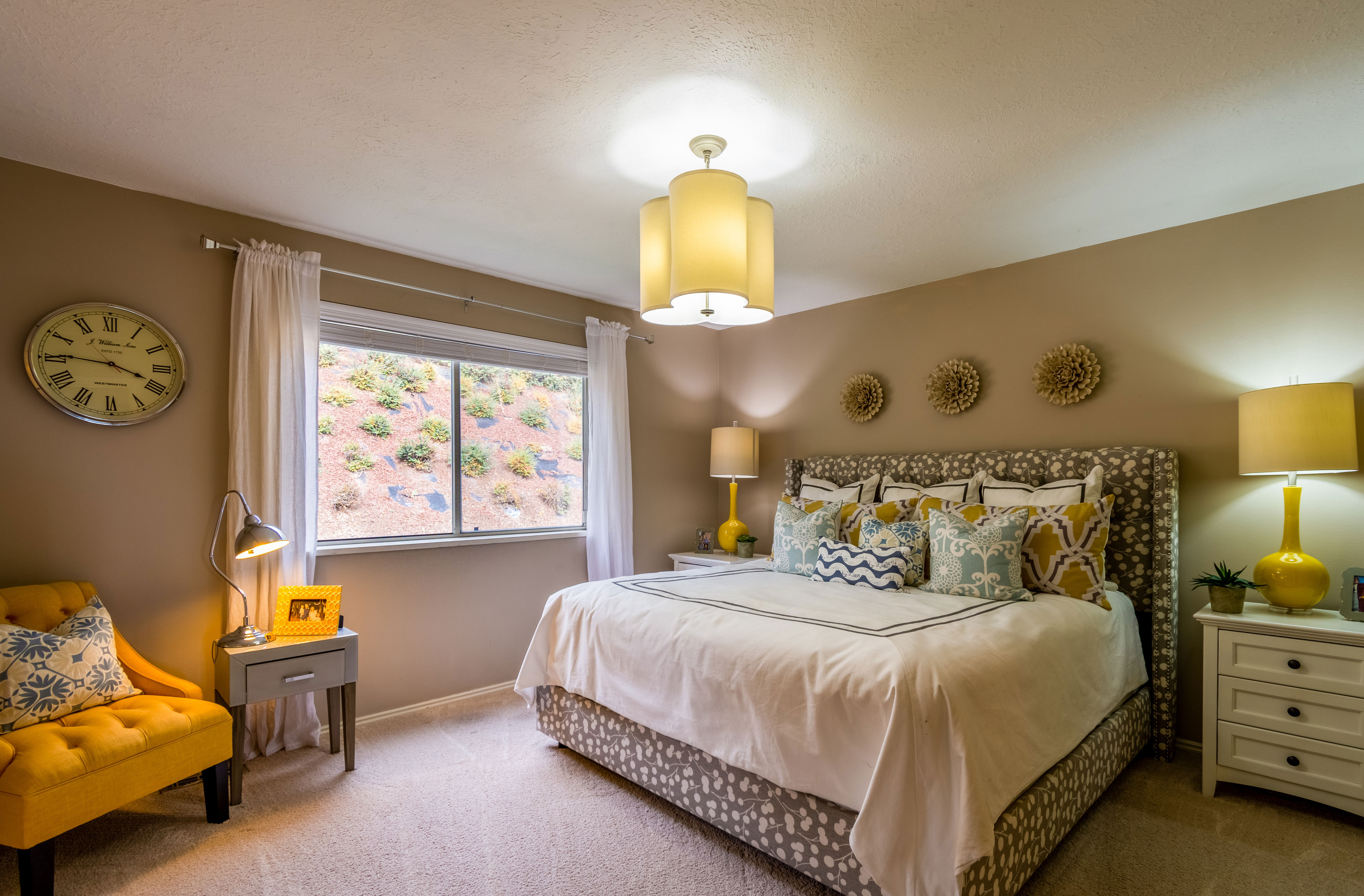 Taupe-colored bedroom with decorative pillows on the bed, a wall clock, and ambient lighting enhancing the cozy atmosphere.