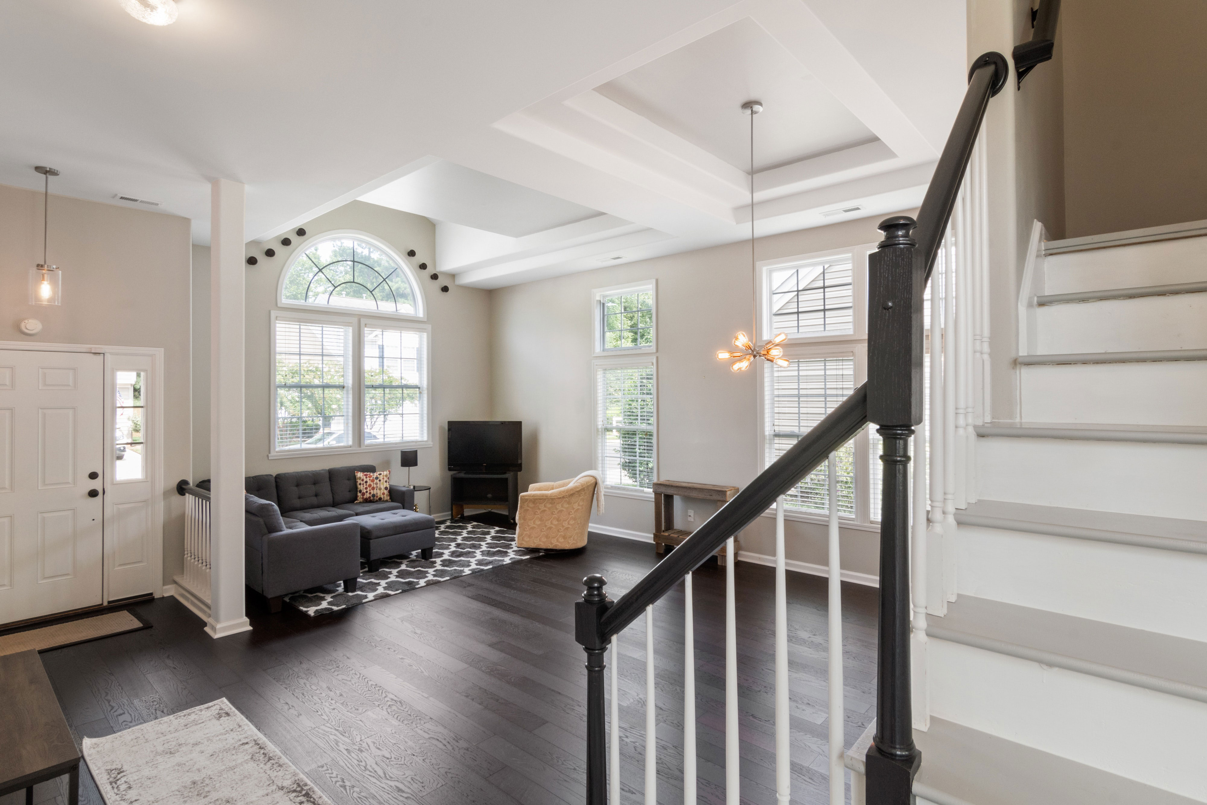 Bright living room with taupe walls, modern furniture, and large windows, featuring a stylish chandelier and staircase.