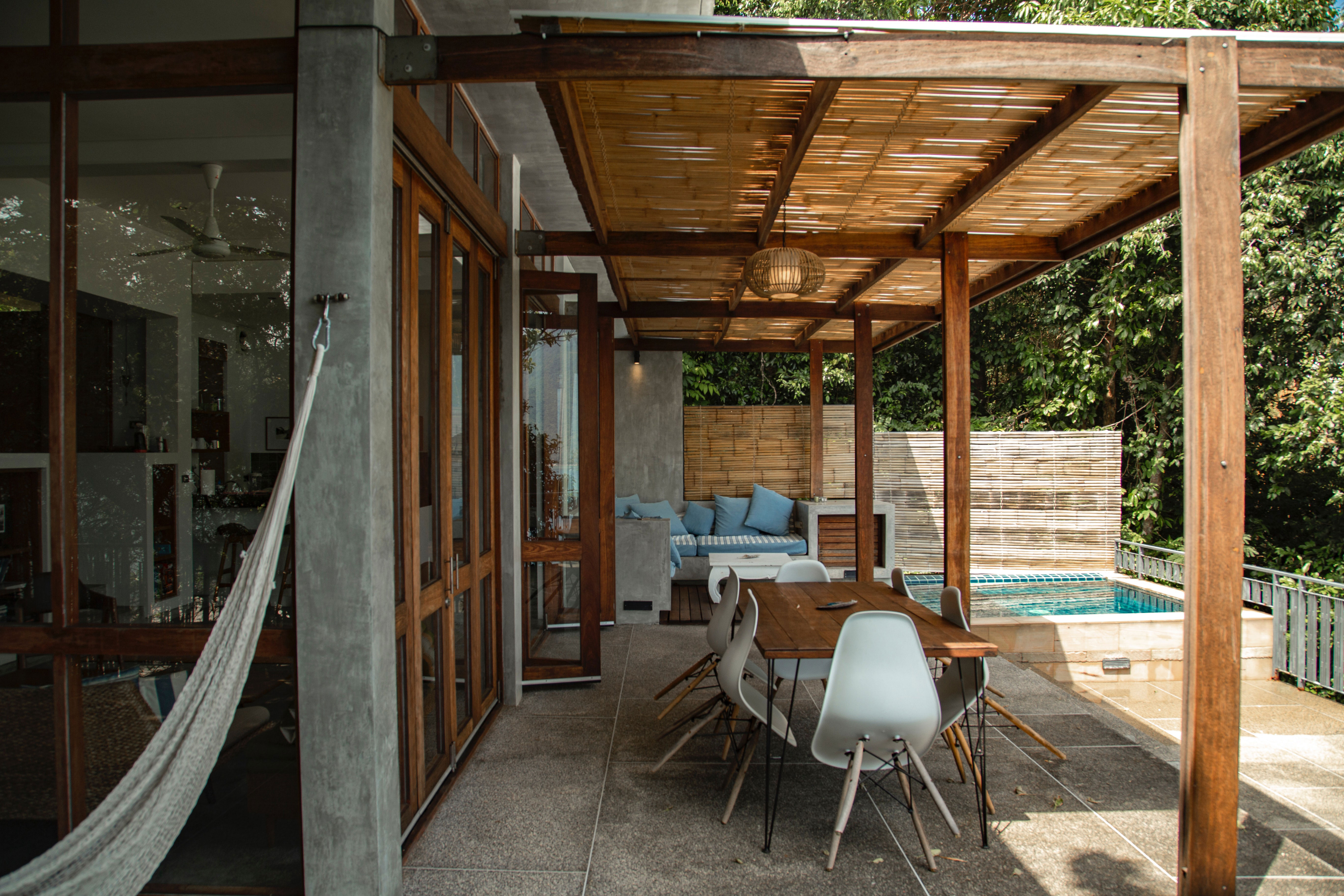 Taupe-themed patio with wooden pergola, dining table, chairs, and cozy seating area by a pool.
