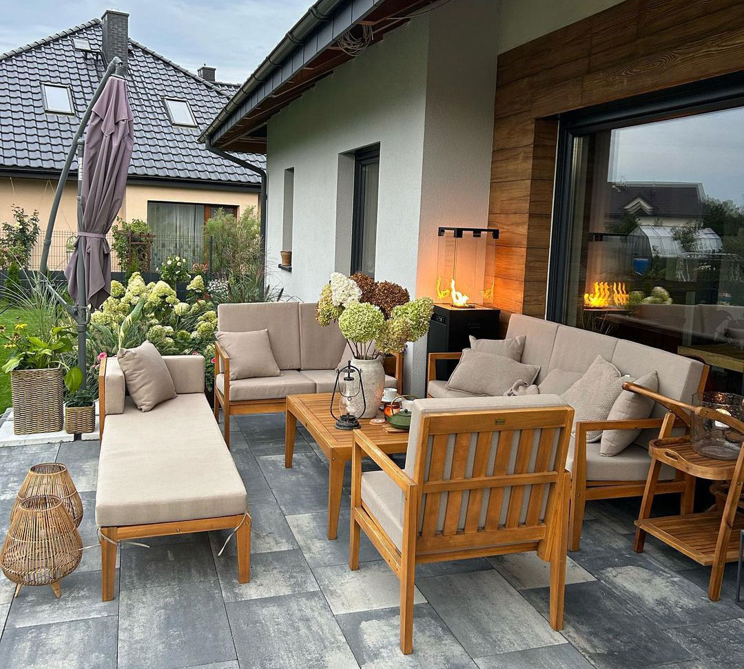 Outdoor patio with taupe furniture, featuring wooden chairs and a sofa, surrounded by greenery and decorative plants.